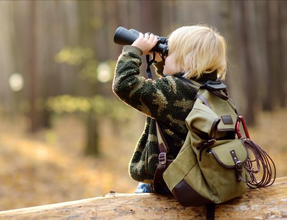 Ein kleiner blonder Junge sitzt mit einem Fernglas auf einem Baumstamm